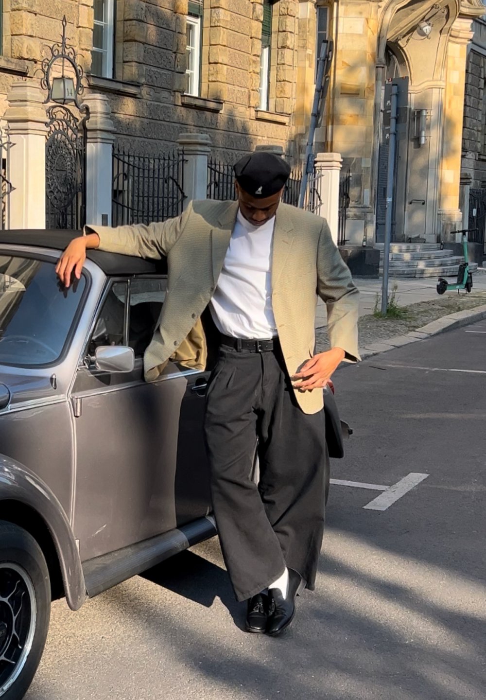 Osoba u bež kariranom saku i bijeloj košulji naslonjena je na sivu vintage automobil, noseći crne hlače i cipele s bijelim čarapama.
