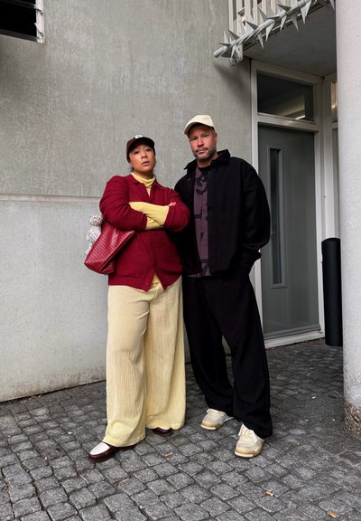 Dos personas están de pie en una calle empedrada junto a un edificio; una lleva una chaqueta roja y pantalones amarillos, la otra lleva una chaqueta y pantalones negros.