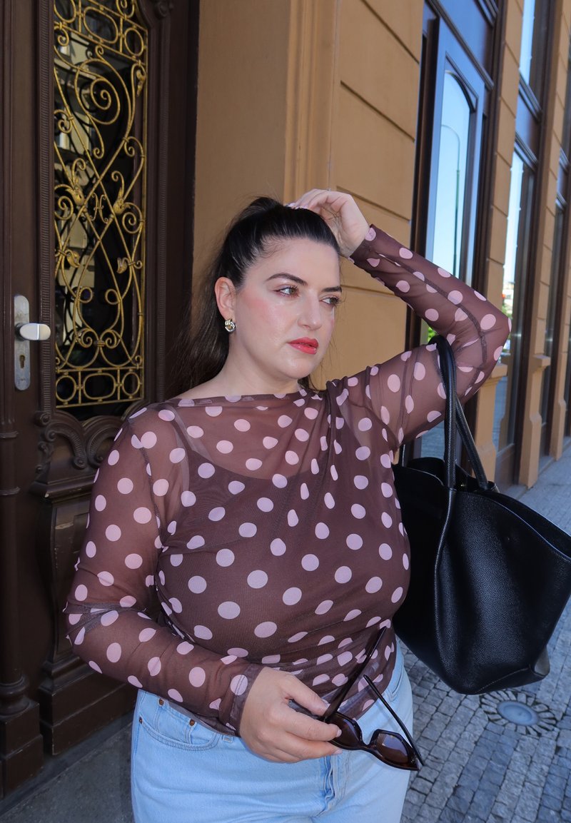 Femme aux cheveux foncés portant un haut transparent à pois et un jean clair, tenant des lunettes de soleil et un sac cabas noir, debout sur un trottoir en ville.