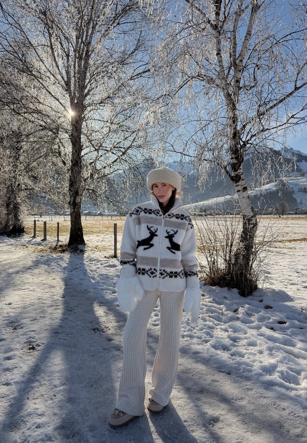 Kobieta w zimowych ubraniach, z kurtką z motywem renifera i rękawiczkami, stoi na zaśnieżonej ścieżce, słońce przebija się przez oszronione drzewa, w tle góry.