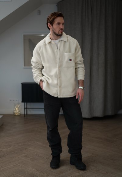 Camisa chaqueta de forro polar crema con un bolsillo frontal y cierre de botones, combinada con pantalones oscuros y botas negras, en un entorno interior moderno.