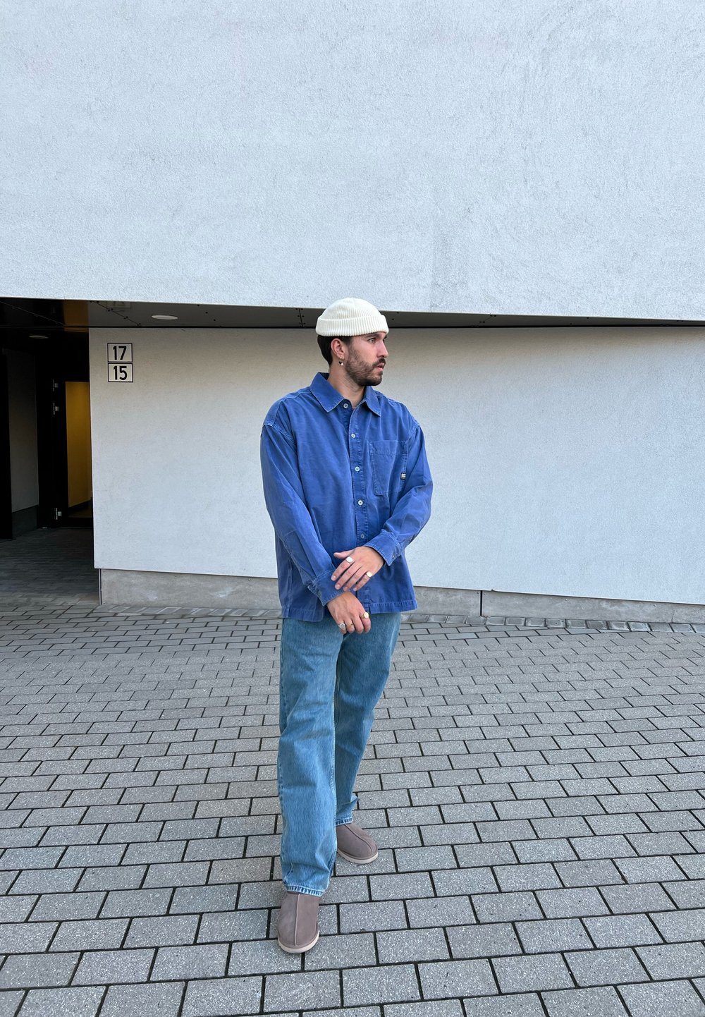 Camisa azul de botões, jeans azul claro e botas cinzas. Acessórios incluem um gorro branco de malha e um anel. Fundo simples com pavimentos.