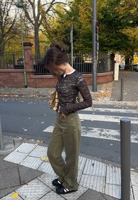 Crna čipkasta bluza dugih rukava, maslinasto zelene hlače širokih nogavica, crne sandale s bijelim prugama. Vanjski urbani prostor s drvećem i pločnikom.