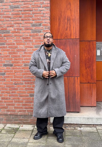 Hombre con el cabello trenzado y gafas, vistiendo un abrigo largo gris, pantalones negros y zapatos negros, de pie frente a una pared de ladrillos y paneles de madera.