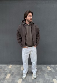 Man with hands in pockets wearing brown jacket, brown hoodie, white trousers, and white trainers, standing against a grey wall.