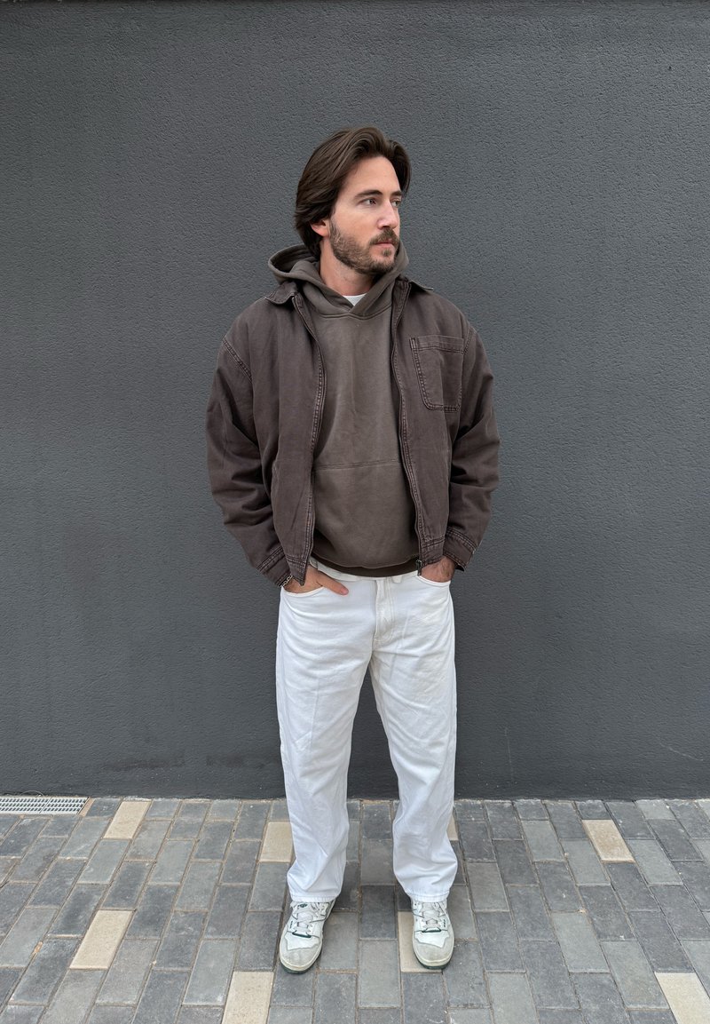 Man with hands in pockets wearing a brown jacket, brown hoodie, white trousers, and white trainers, standing against a grey wall.