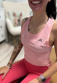 Top desportivo rosa com design perforado, emparelhado com leggings rosas combinando. Apresenta o logo da Adidas no peito e pulseiras empilhadas no pulso.