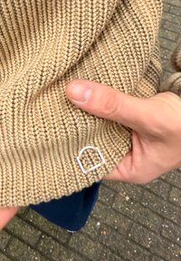 Megztinis megztas iš smėlio spalvos siūlų, su turinčia gyslų tekstūra, prie apačios puošiamas sidabro spalvos stačiakampio formos metaliniu akcentu. Ranka laiko audinį.