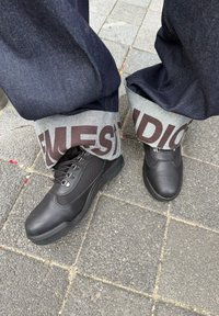 Scarpe da trekking nere indossate con jeans di un blu scuro risvoltati per rivelare un audace tessuto bianco con grandi lettere marroni su un marciapiede lastricato.