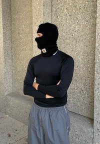 Balaclava de punto negra con el logo de Carhartt, usada con una camiseta de manga larga negra y pantalones cortos grises con bolsillos laterales. Fondo de concreto texturizado.
