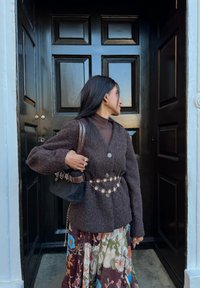 Cardigan marron texturé avec de gros boutons, porté sur une jupe florale à motifs. Accessoirisé d'un petit sac noir et d'une ceinture décorative.