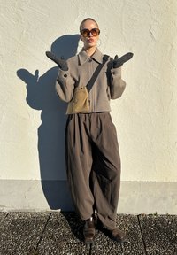 Veste marron oversize avec grand col, pantalon ample marron, gants gris, lunettes de soleil et petit sac crossbody beige clair devant un mur blanc.