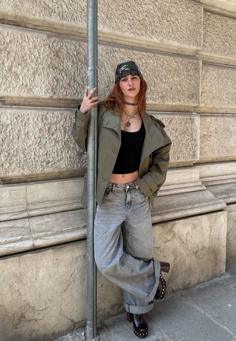 Νεαρό άτομο με κόκκινα μαλλιά, με καμουφλαρισμένο μαντήλι στο κεφάλι, ελαιρώδες μπουφάν, μαύρο κομψό τοπ, φαρδιά τζιν και ξύλινα παπούτσια, στηρίζεται σε κολώνα δίπλα σε μια υφή πέτρινος τοίχο.