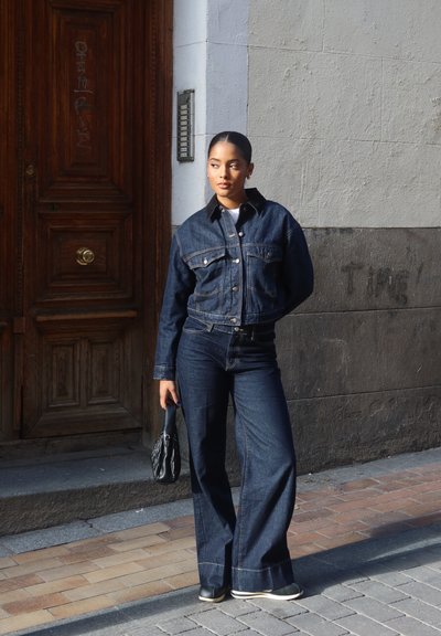 Chaqueta de mezclilla oscura con bolsillos en el pecho y botones, combinada con jeans anchos oscuros. Accesorios incluyen un pequeño bolso negro. Fondo de puerta de madera.