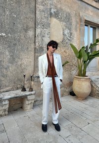 Uomo in abito bianco, camicia marrone con una lunga sciarpa, occhiali da sole neri e scarpe nere, che si trova di fronte a un muro di pietra logorato con una grande pianta in vaso nelle vicinanze.