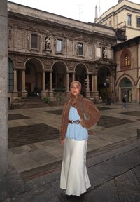 Eine Frau steht auf einem Platz und trägt eine braune Kunstpelzjacke, ein hellblaues Hemd und einen langen weißen Satinenrock, ergänzt durch eine grüne Tasche und einen Gürtel.