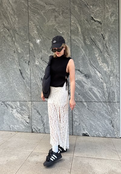 Top negro de canalé sin mangas, falda maxi blanca de encaje con aberturas delanteras, gorra negra, zapatillas negras con rayas blancas y calcetines blancos.