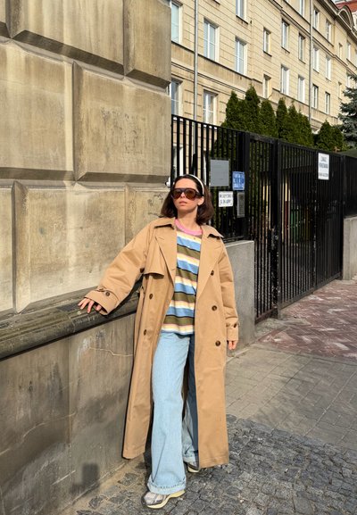 Mujer con gabardina beige, camisa de rayas, vaqueros azul claro y gafas de sol apoyada contra una pared de piedra cerca de una verja metálica negra en un entorno urbano.