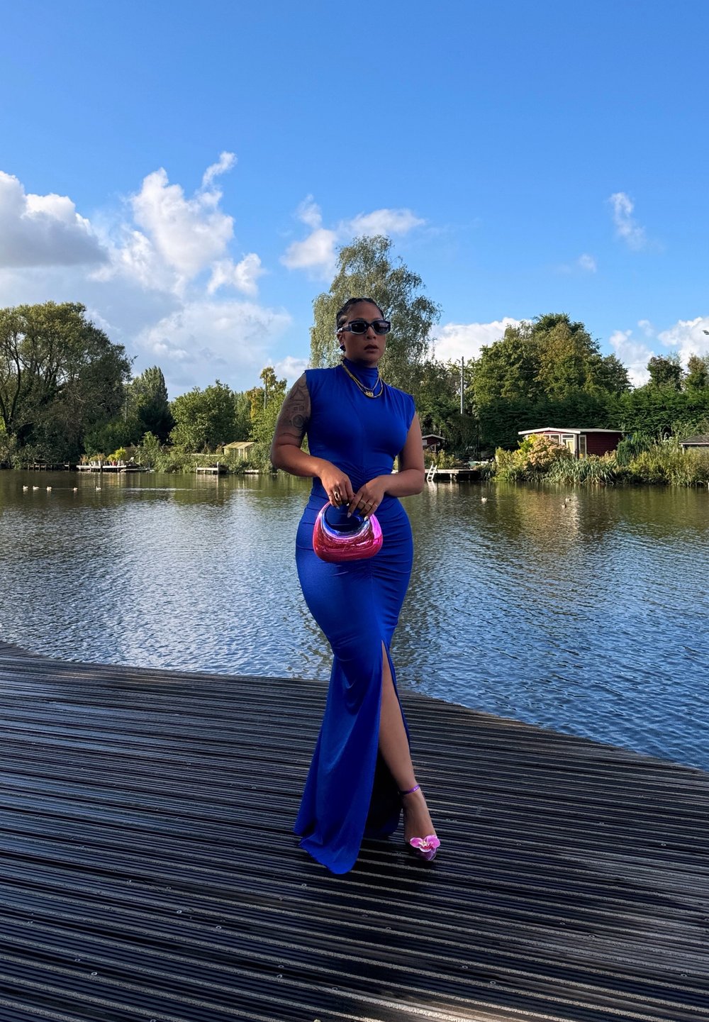 Robe ajustée bleue avec une fente sur le côté, en tissu lisse, accessoires dorés et un sac à main rose. Se tenant devant un arrière-plan aquatique serein.
