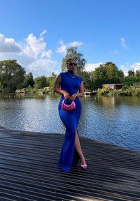 Robe ajustée bleue avec une fente sur le côté, en tissu lisse, accessoires dorés et un sac à main rose. Se tenant devant un arrière-plan aquatique serein.