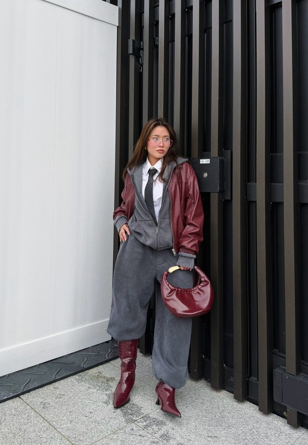 Young woman wearing a burgundy jacket, grey hoodie, loose grey trousers, and burgundy boots, holding a matching handbag, standing against a black fence.