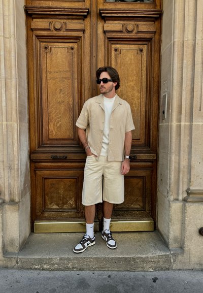 Camisa beige de manga corta, camiseta blanca, pantalones cortos beige y zapatillas negras con detalles blancos, de pie frente a una puerta de madera.