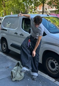 Calças de ganga largas com bainhas desfarrapadas, combinadas com uma t-shirt cinza e sapatos pretos. Saco de lona verde no chão ao lado de uma carrinha branca estacionada.