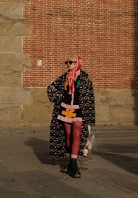 Persona che indossa un cappotto lungo a fantasia, un cappello colorato lavorato a maglia, collant rossi, stivali neri e porta una borsa argento, in piedi su una strada contro un muro di mattoni.