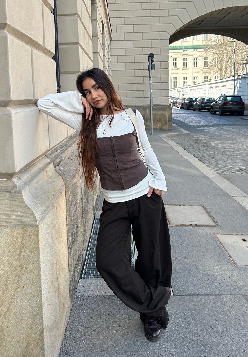 Femme aux longs cheveux appuyée contre un mur en pierre, portant un haut blanc à manches longues, un corset marron, un pantalon large noir et des chaussures noires dans une rue en ville.