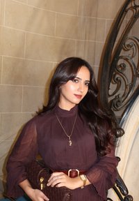 Brązowa pikowana torebka na łańcuszku ze złotą klamrą, noszona z ciemnofioletową bluzką z rozkloszowanymi rękawami oraz naszyjnikiem z zawieszką. Miękkie oświetlenie wewnętrzne.