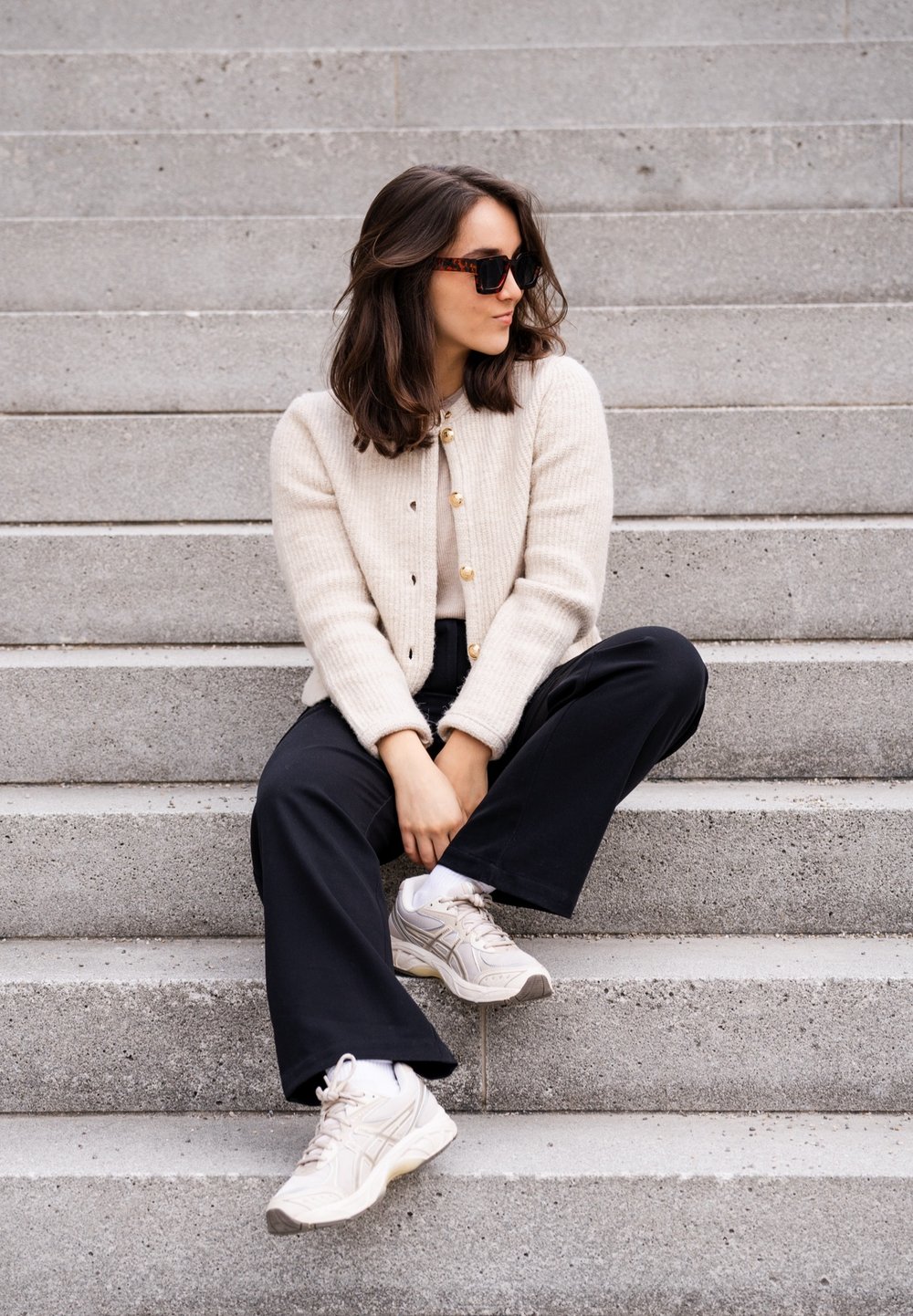 Beżowy, prążkowany kardigan z złotymi guzikami, czarne spodnie typu wide-leg i jasne sneakersy. Modelka siedzi na szarych kamiennych schodach w swobodnej pozie.