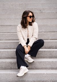 Beżowy, prążkowany kardigan z złotymi guzikami, czarne spodnie typu wide-leg i jasne sneakersy. Modelka siedzi na szarych kamiennych schodach w swobodnej pozie.