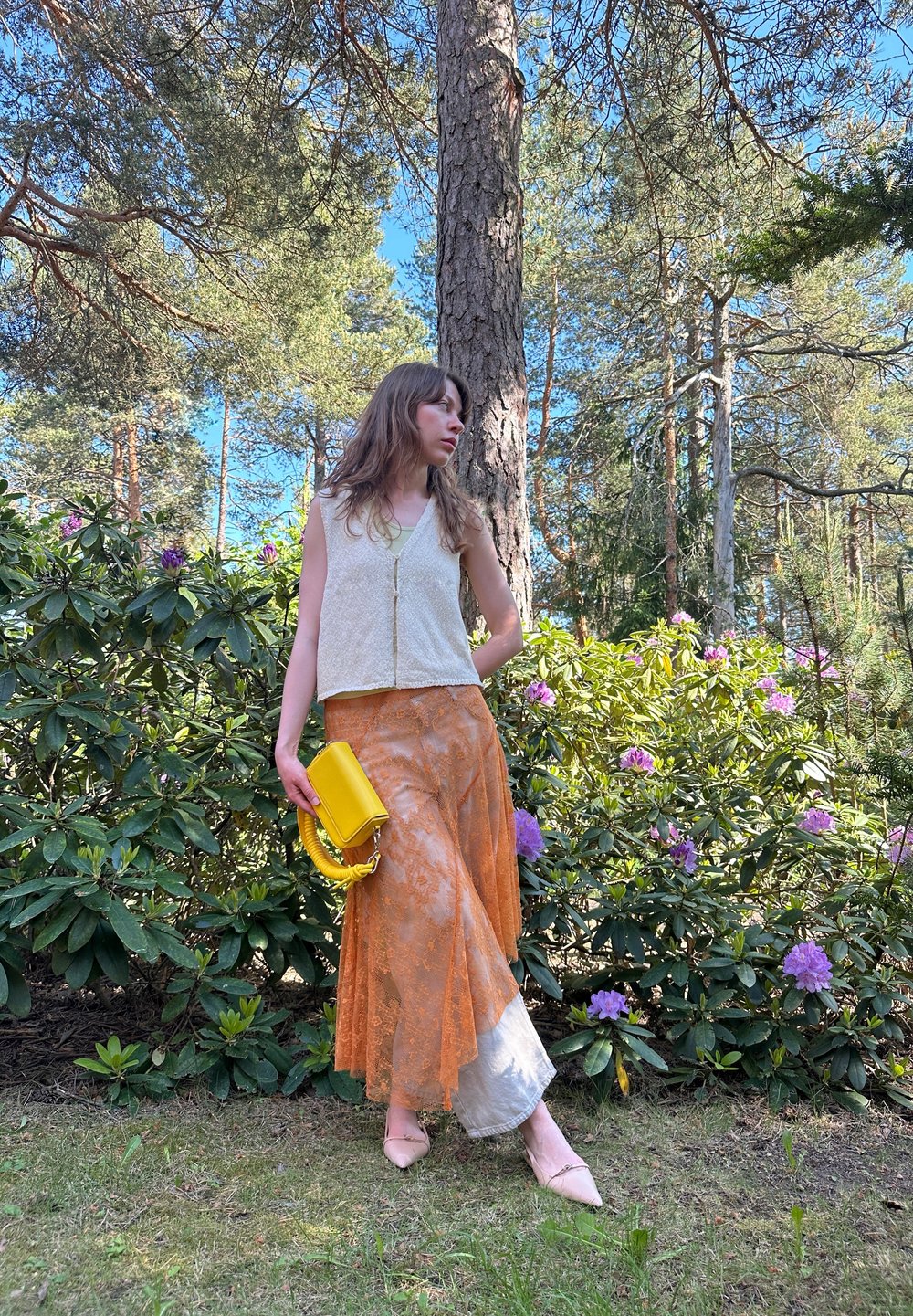 Beige kort topp, oransje blonde skjørt med satengkant, og en gul håndveske med håndtak, satt mot grønne blader og blomster.