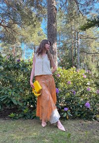 Beige kort topp, oransje blonde skjørt med satengkant, og en gul håndveske med håndtak, satt mot grønne blader og blomster.