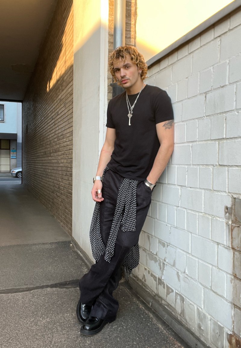 Jeune homme aux cheveux bouclés portant un t-shirt noir et un pantalon, appuyé contre un mur en briques blanches dans une ruelle urbaine au coucher du soleil.