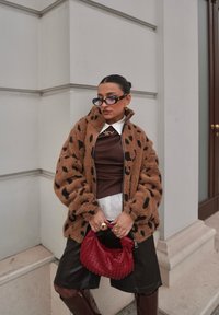 Veste texturée marron à pois noirs, haut marron, chemise blanche, short noir, tenant un sac tissé rouge. Bottes marron montant jusqu'aux genoux complètent le look.