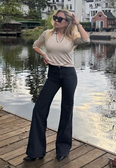 Suéter de punto beige con escote en V, acompañado de jeans de campana negros de cintura alta y tacones puntiagudos negros, situado junto a una superficie de agua reflectante.