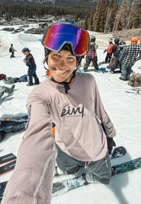 Snowboarder îmbrăcat cu o bluză roz deschis cu mânecă lungă și pantaloni negri, purtând ochelari de protecție colorați, stând pe zăpadă lângă alți snowboarderi.