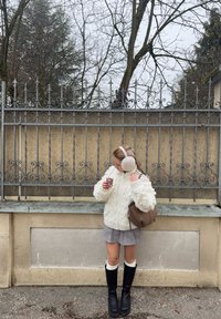 Pakse valge soonikuleeritud kampsun, hall seelik, põlveni ulatuvad mustad saapad, helepruun käekott, hoiab tassikest, kasuka kõrvarätikud, rauast aiaga taustal.