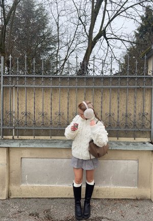 Suéter blanco de punto grueso con cable, falda gris, botas negras hasta la rodilla, bolso marrón claro, sosteniendo una taza, con orejeras de piel, frente a una cerca de hierro forjado.