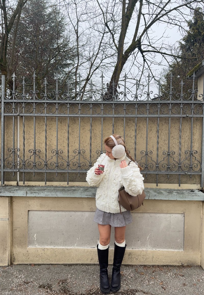 Pull en maille blanche épaisse, jupe grise, bottes noires montantes, sac à main marron clair, tenant une tasse, avec des cache-oreilles en fourrure, devant une grille en fer forgé.