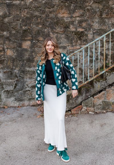 Cárdigan a rayas en azul verdosa y blanco, top negro, falda blanca y zapatillas azul verdosa. Sosteniendo un bolso negro. Muro de piedra de fondo.