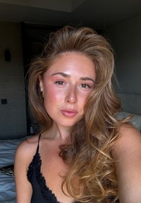 Ritratto di una persona con lunghi capelli ondulati castani e tratti delicati, che indossa un top in pizzo nero, con una carnagione naturale e un trucco leggero.