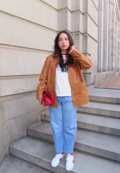 Mujer con chaqueta marrón, camiseta blanca con gráfico, vaqueros azul claro y zapatillas blancas que sostiene un bolso rojo y está parada en unos escalones de piedra al aire libre.
