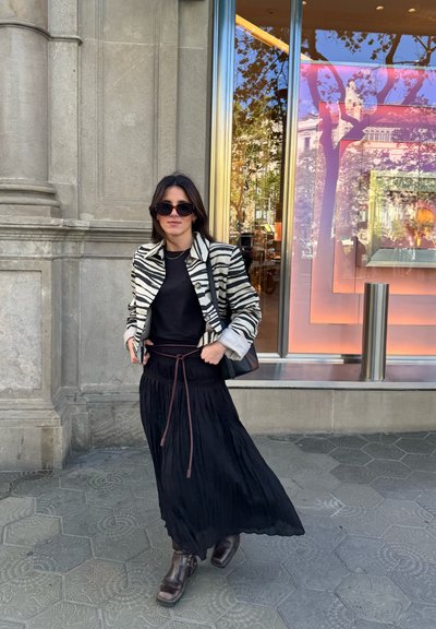 Chaqueta a rayas de cebra, top negro, falda plisada negra con cuerda de ajuste marrón y botas de tacón marrón, frente a una tienda con escaparate reflectante.