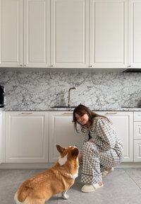 Femme en pyjama à carreaux accroupie pour caresser un chien corgi dans une cuisine moderne avec des armoires blanches et une crédence en marbre.