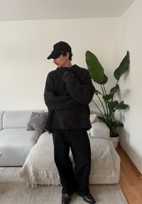 Pull noir en maille épaisse avec un col montant, assorti à un pantalon noir ample. Le décor comprend un canapé clair et une plante d'intérieur.