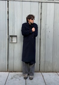 Manteau long bleu marine en tissu épais, avec de gros boutons, porté sur un pantalon gris ample et des chaussures foncées. Debout devant un mur en métal ondulé.