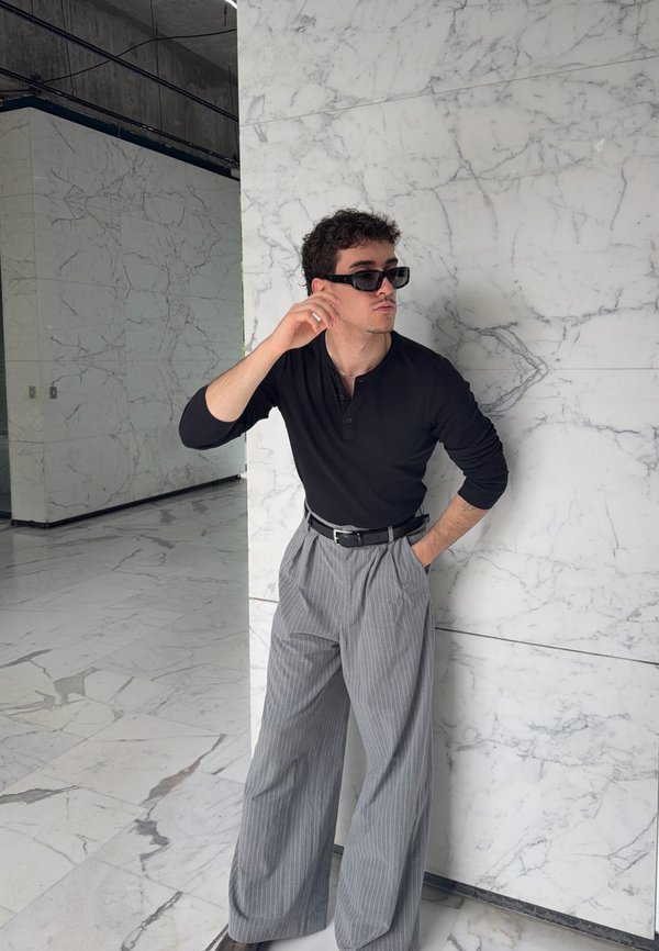 Mann trägt schwarzes Langarmshirt, graue Nadelstreifenhose mit weitem Bein und schwarze Sonnenbrille und posiert vor einer weißen Marmorwand im Innenraum.