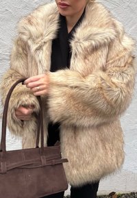 Valge beeži tooni kunstkarusnahast mantel, pehme tekstuuriga, lai krae ja ülisuurte varrukatega. Kõrgepruun nobenahast käekott hoitakse ühes käes.